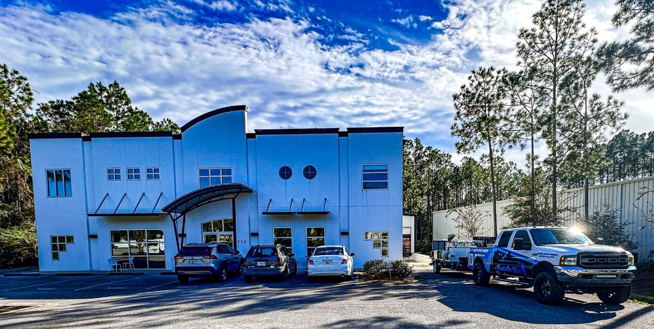 Commercial building with a curved architectural design, featuring a building washing truck parked in front. The sky is blue with scattered clouds, and the building is surrounded by trees.