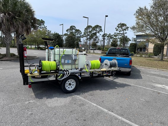 Blue pickup truck towing a trailer with a water tank and hose reels for parking lot and garage cleaning services. The trailer is parked in a parking lot with a Verizon store in the background.