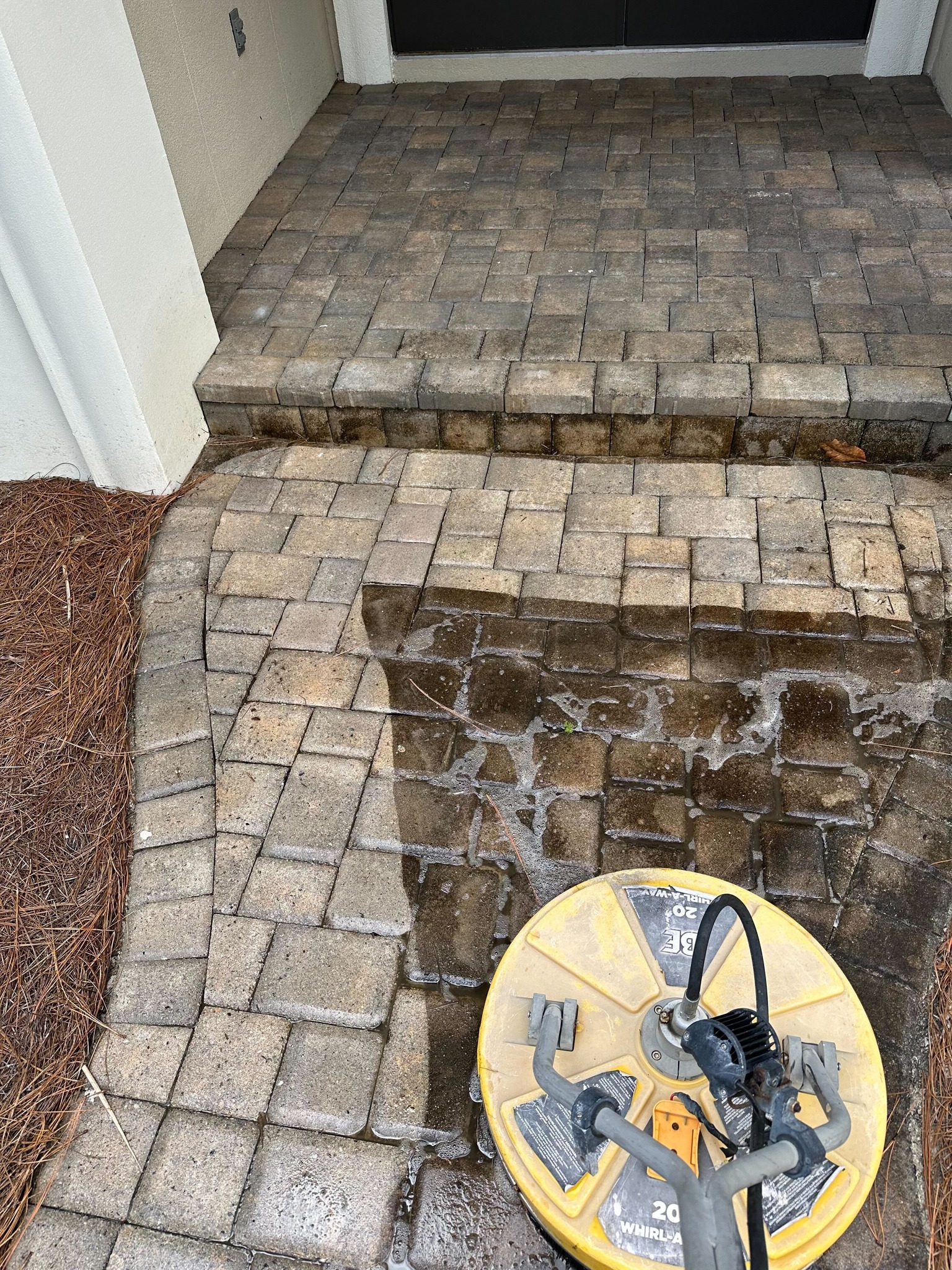 Callaway, FL pressure washing in progress on a brick paver walkway. A yellow surface cleaner is removing grime, revealing the clean pavers beneath.