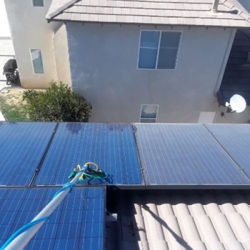 Close-up shows a worker using a brush on a pole to perform solar panel cleaning on rooftop panels. A house and lawn are visible in the background.