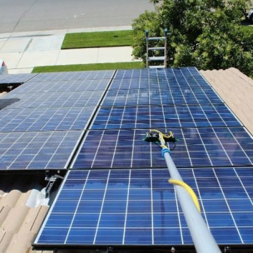 Cleaning solar panels on a residential rooftop. A long-handled brush is used to gently scrub the blue solar panels. A ladder is visible, with green lawn and a suburban street in the background.