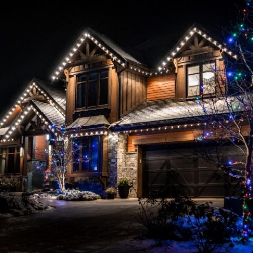 Beautiful home adorned with white Christmas lights along the roofline, and a tree with colorful lights in the yard. A professional Christmas light installation creates a festive, warm glow on a winter night.