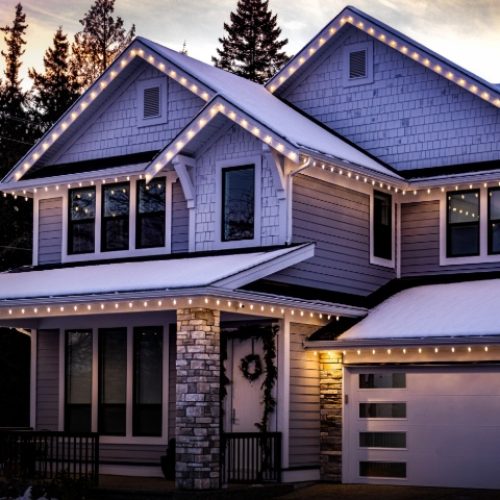 A beautiful two-story house is decorated with warm white Christmas lights along the roofline and porch, set against a snowy winter evening sky.